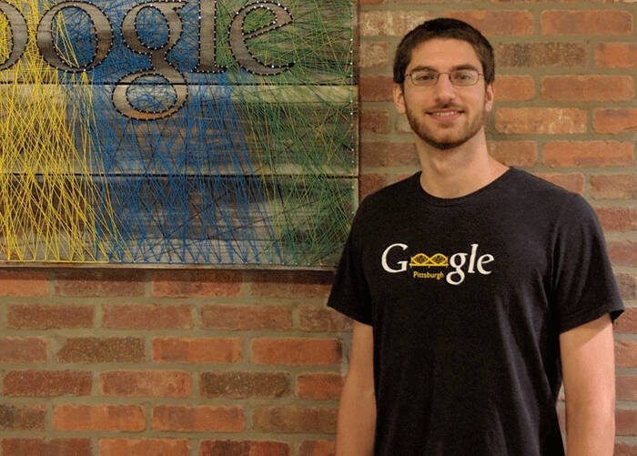 A man wearing glasses and a black 'Google Pittsburgh' t-shirt standing in front of a brick wall with a colorful string art Google logo.